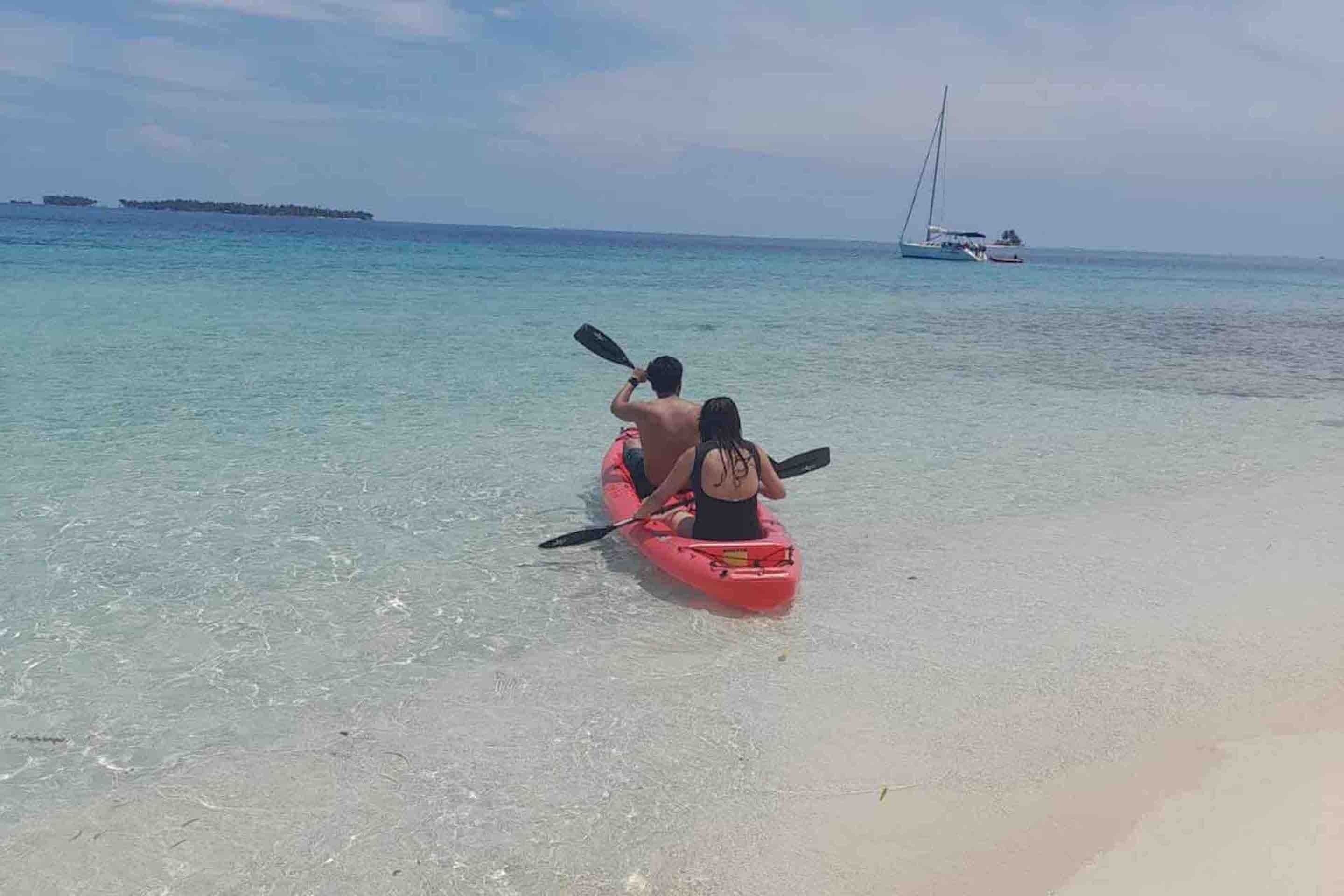 A couple kayaking on a clear beach with a sailboat in the distance.