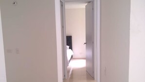 A narrow hallway with white walls and a tiled floor leading to a bedroom with a bed and a nightstand. - Apartamento conjunto Entre Sierras (Valledupar)
