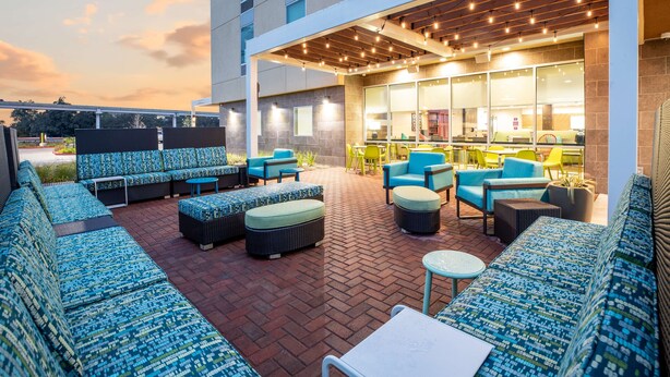 A covered outdoor seating area with sofas and chairs, a brick floor, and a building with large windows in the background. - Home2 Suites by Hilton Houston IAH Airport Beltway 8 (Houston)