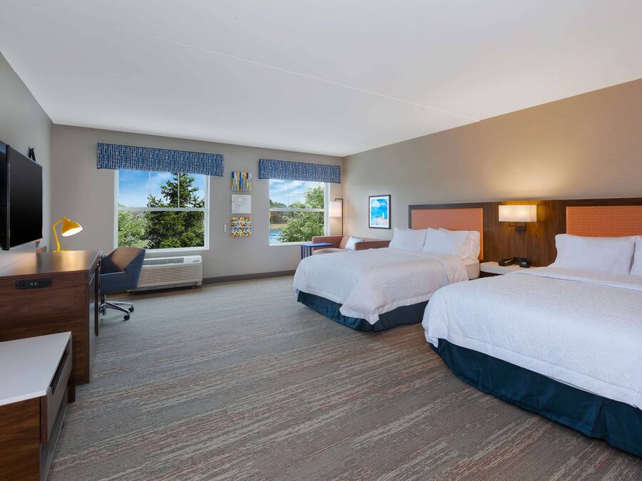 A hotel room with two beds, a desk, a chair, a TV, and a window with a view of greenery.