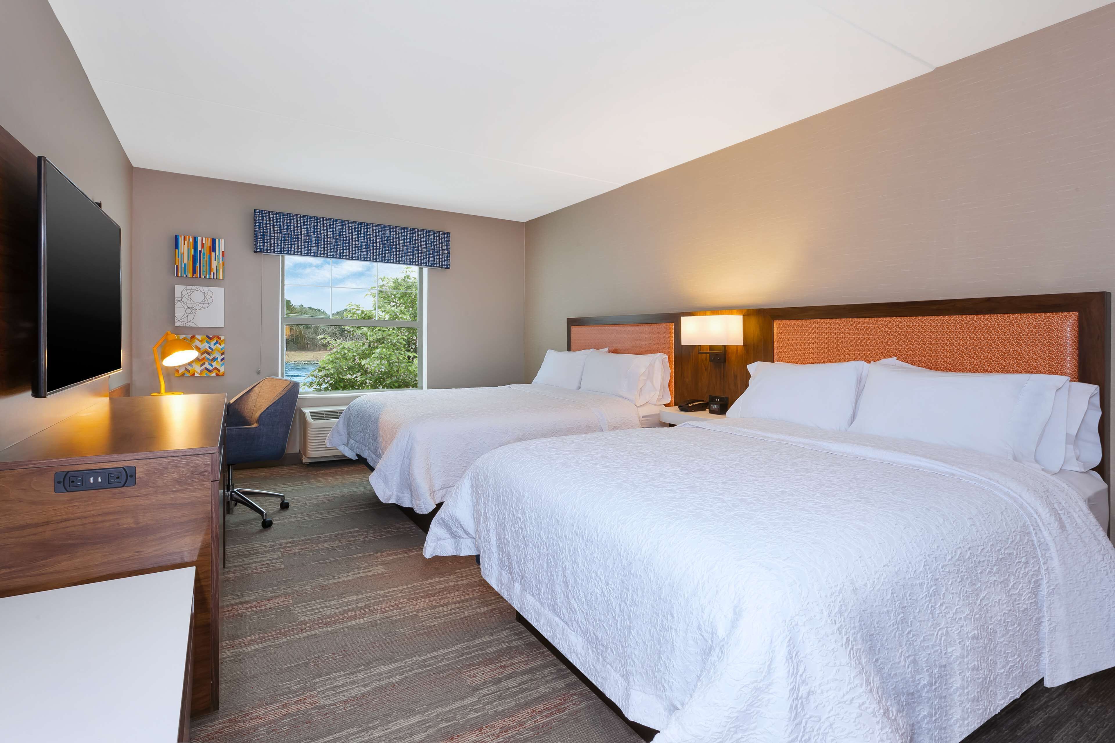 A hotel room with two beds, a desk with a flat-screen TV, a chair, and a window with a view of greenery.