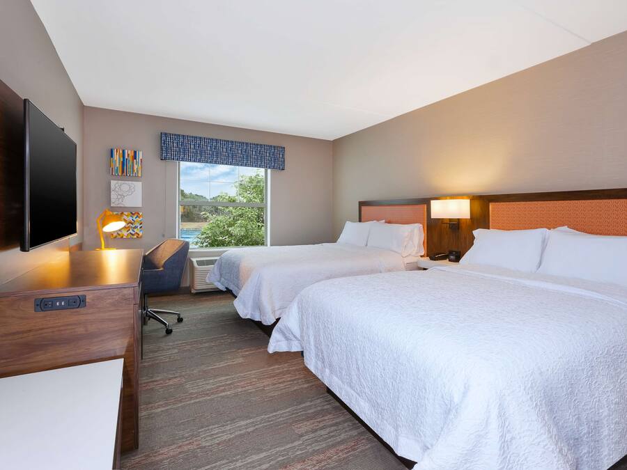A hotel room with two beds, a desk with a flat-screen TV, a chair, and a window with a view of greenery.
