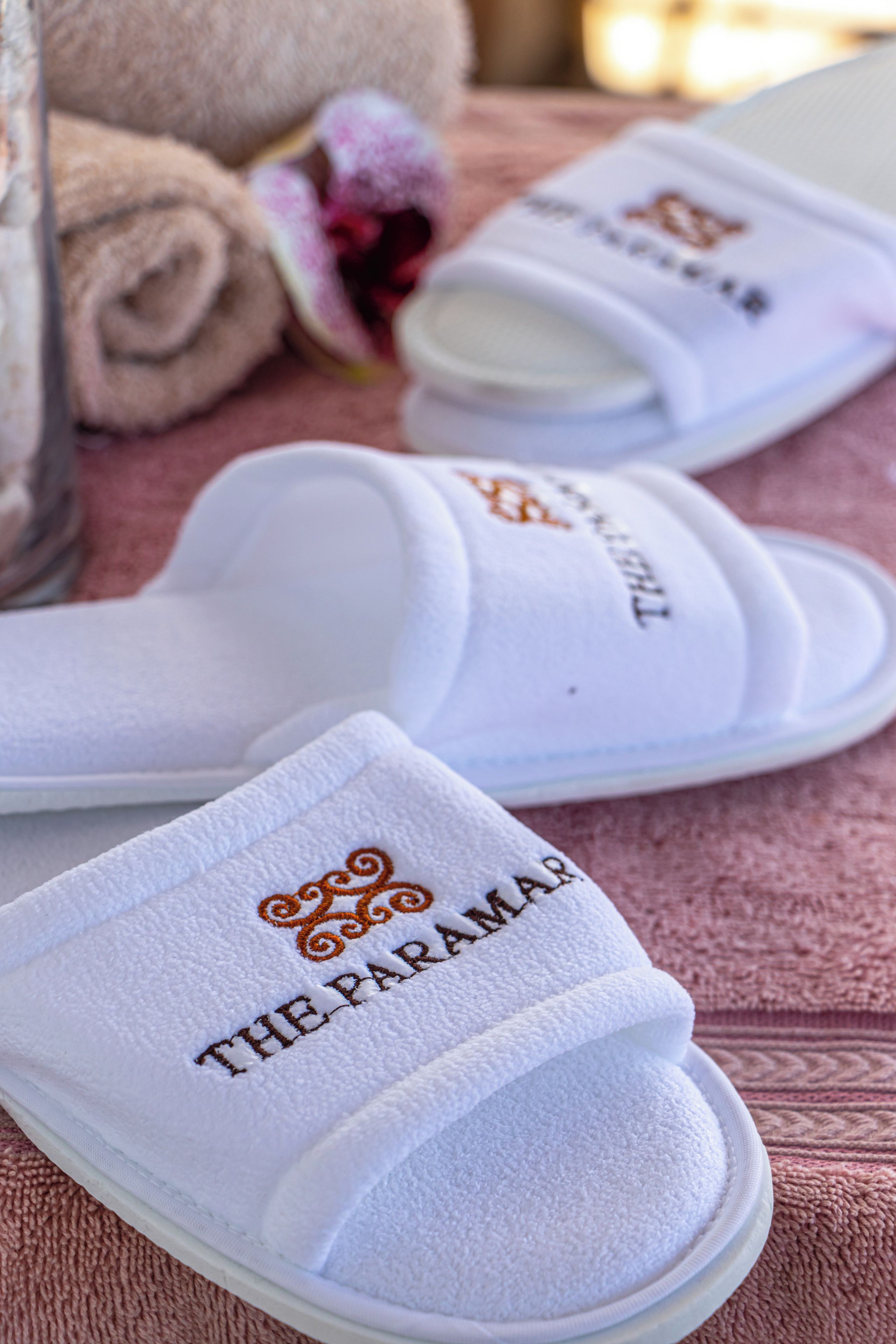 A pair of white slippers with embroidered text and a logo, placed on a textured surface.