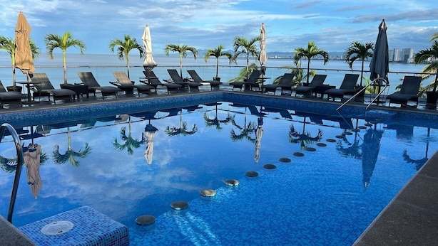 A pool with lounge chairs and umbrellas, surrounded by palm trees, with a view of the sea and buildings in the background. - The Paramar Beachfront Boutique Hotel - Downtown Malecon (Puerto Vallarta)