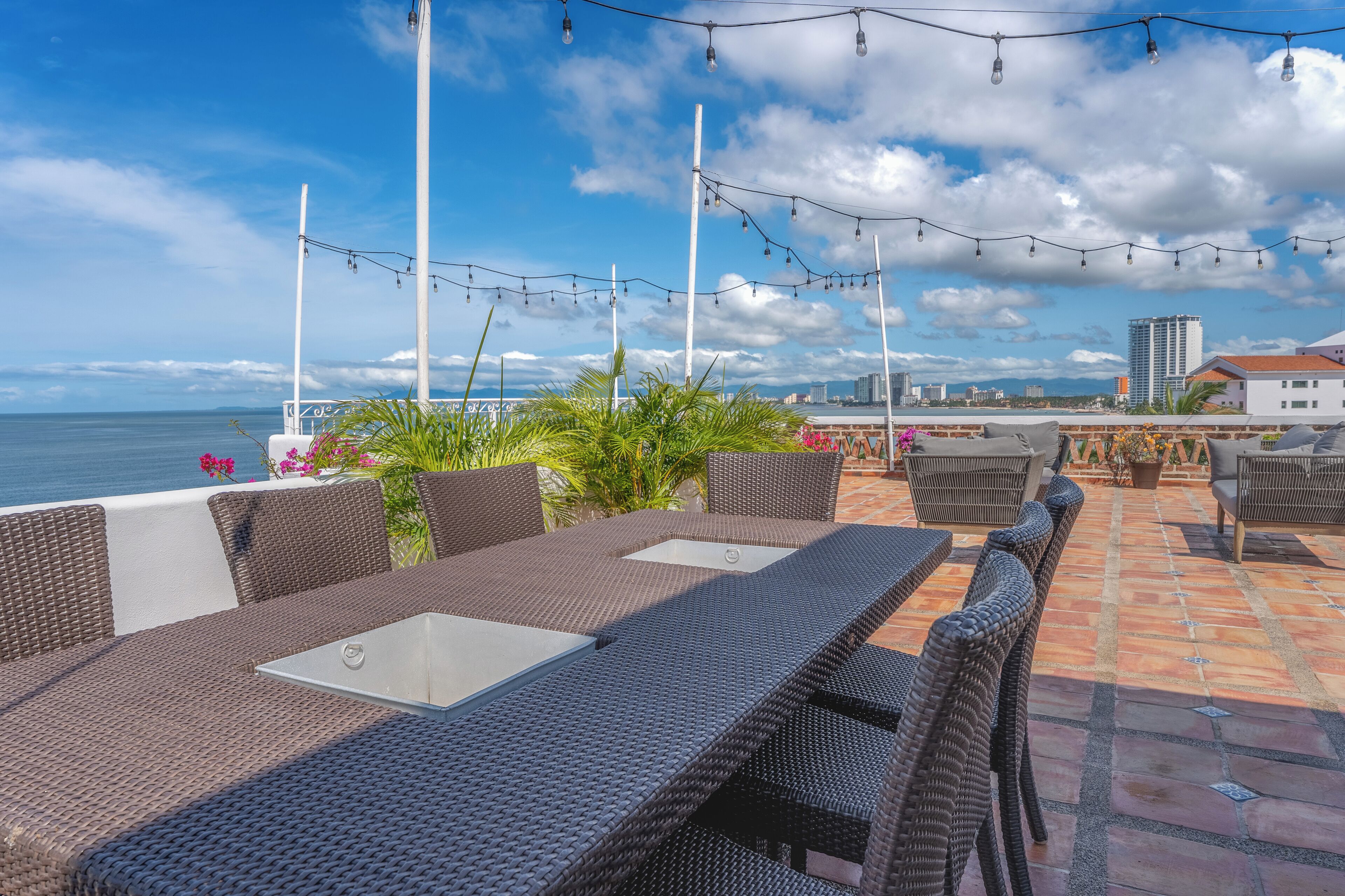 An outdoor patio with wicker furniture, string lights, and a view of the sea.
