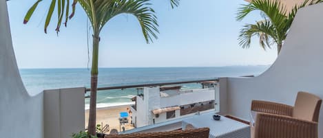 A balcony with wicker furniture, a potted plant, and a view of the beach and ocean.