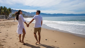 A couple walking hand in hand on a sandy beach with a calm sea and mountains in the background. - The Paramar Beachfront Boutique Hotel - Downtown Malecon (Puerto Vallarta)