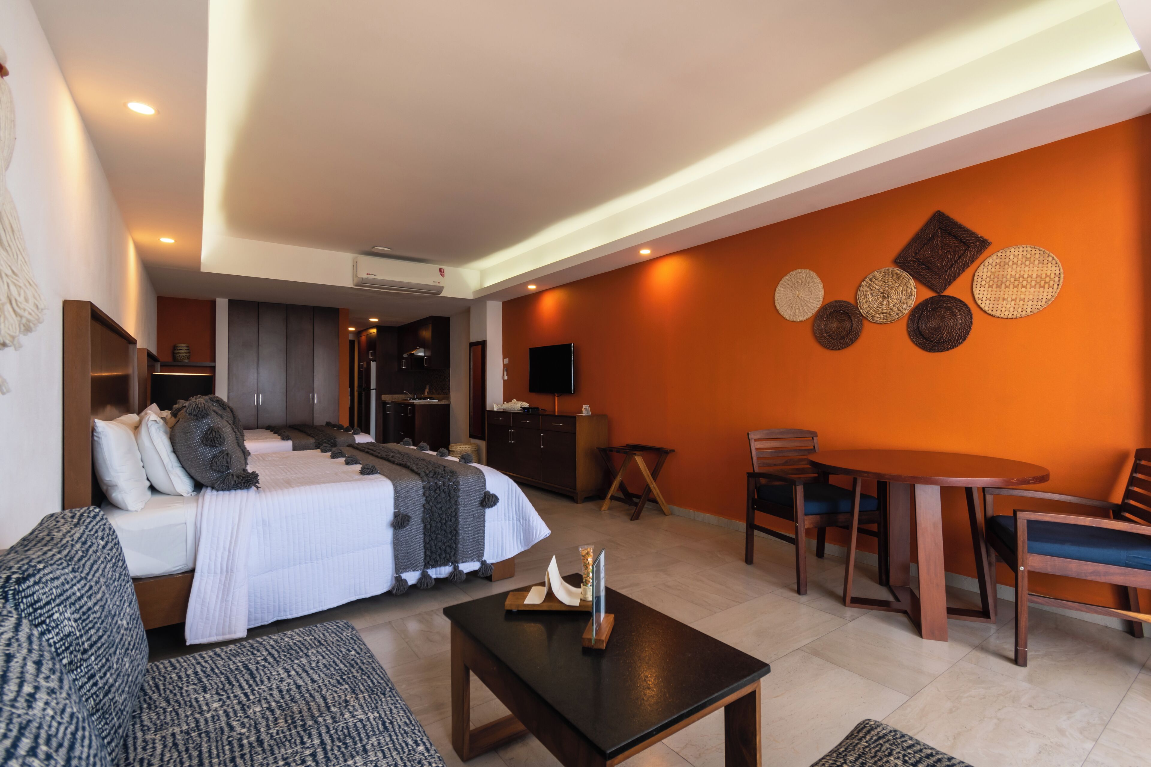 A modern hotel room with a bed, sofa, dining table, and chairs. There is a TV mounted on the wall and a small table with a candle.