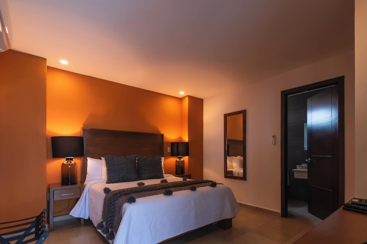 A hotel room with a large bed, two bedside lamps, a nightstand, a wooden headboard, a mirror, and a door leading to a bathroom.