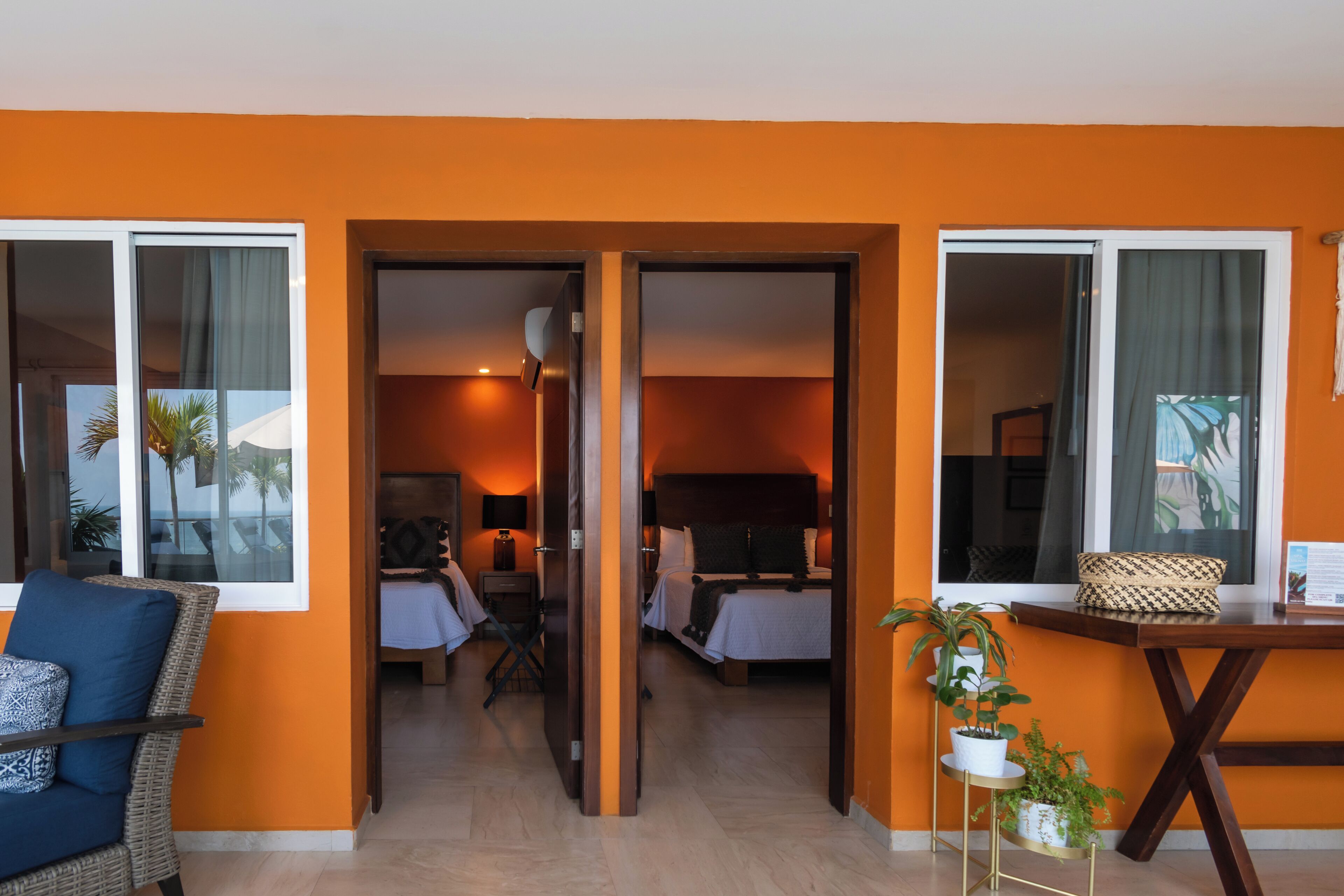 Habitación de hotel con pared naranja, una cama, una silla, una mesita con una planta y vistas a través de una puerta abierta.
