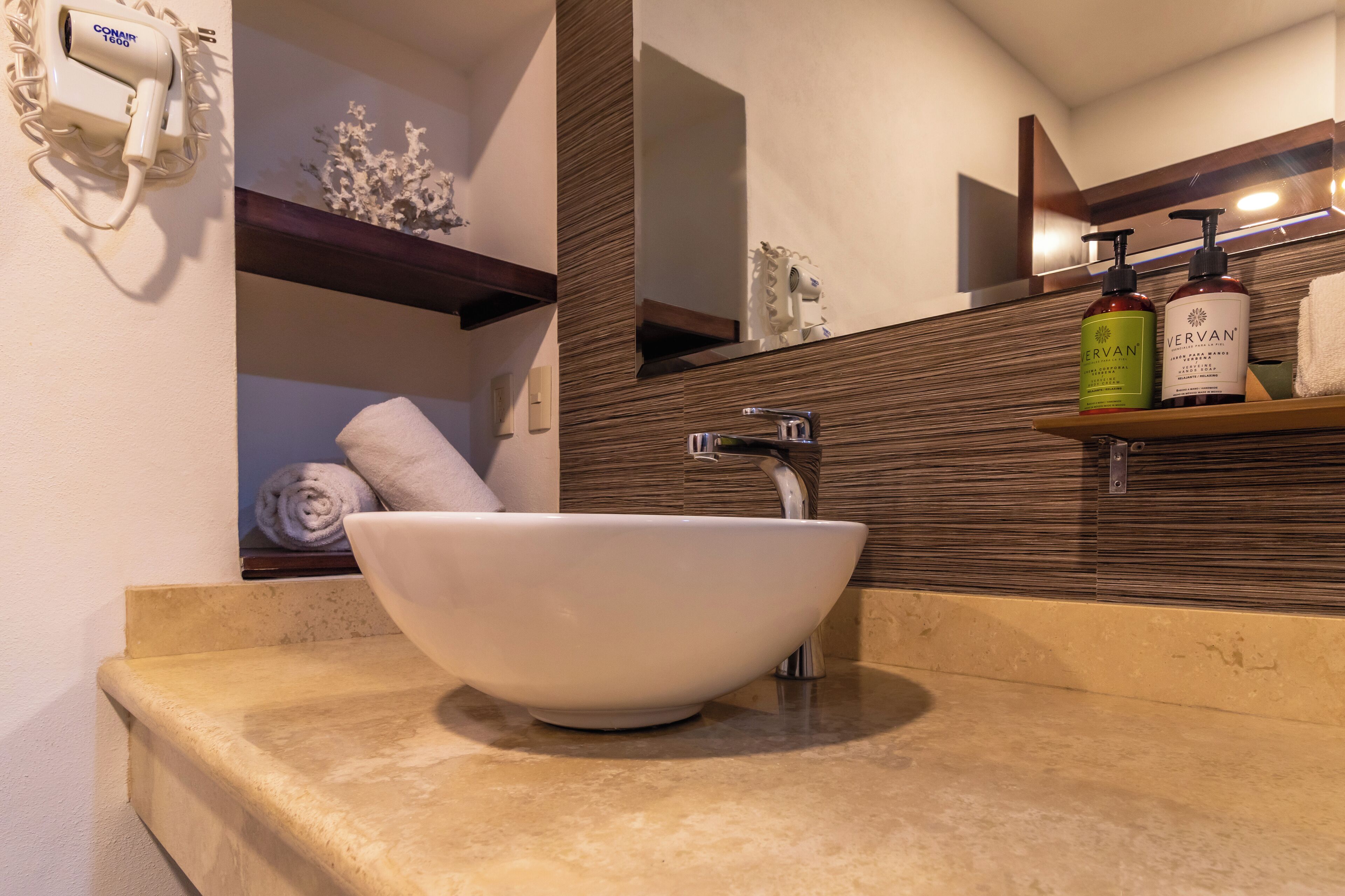 A bathroom with a white vessel sink, a mirror, and a shelf with toiletries.