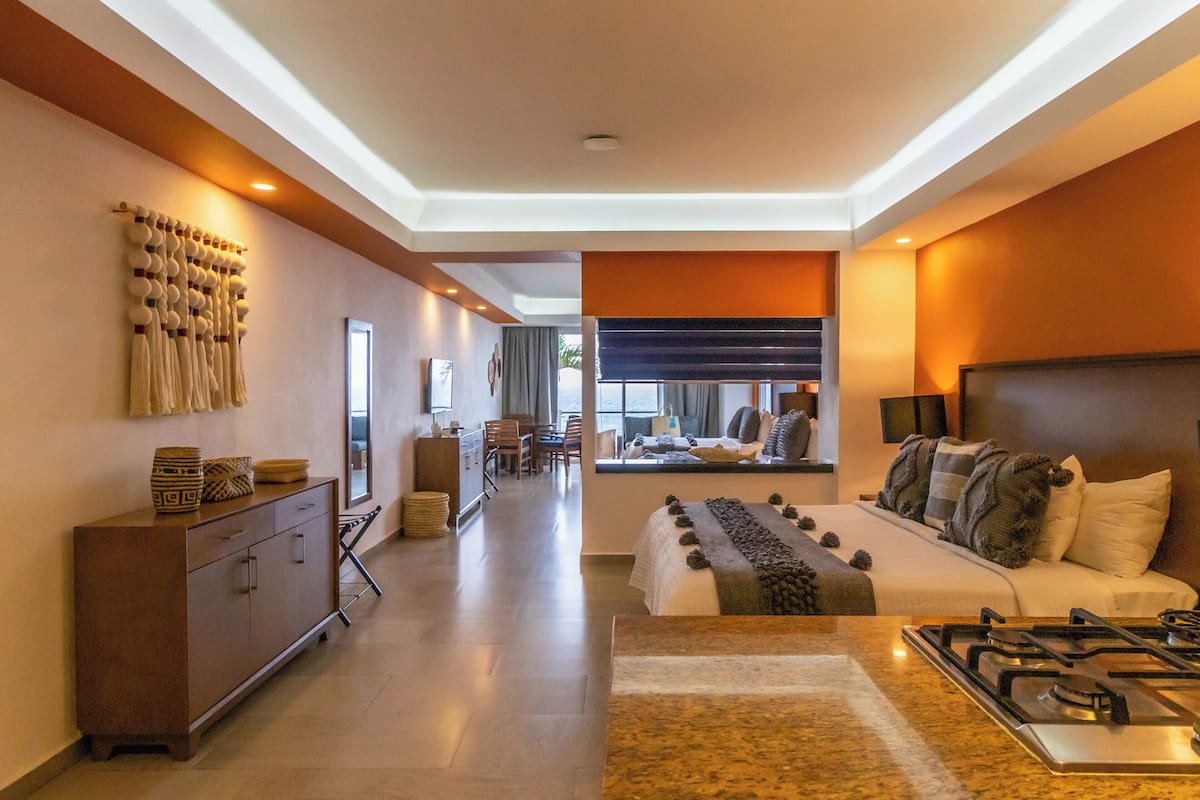 A modern hotel room with a large bed, a kitchen area, and a dining area.