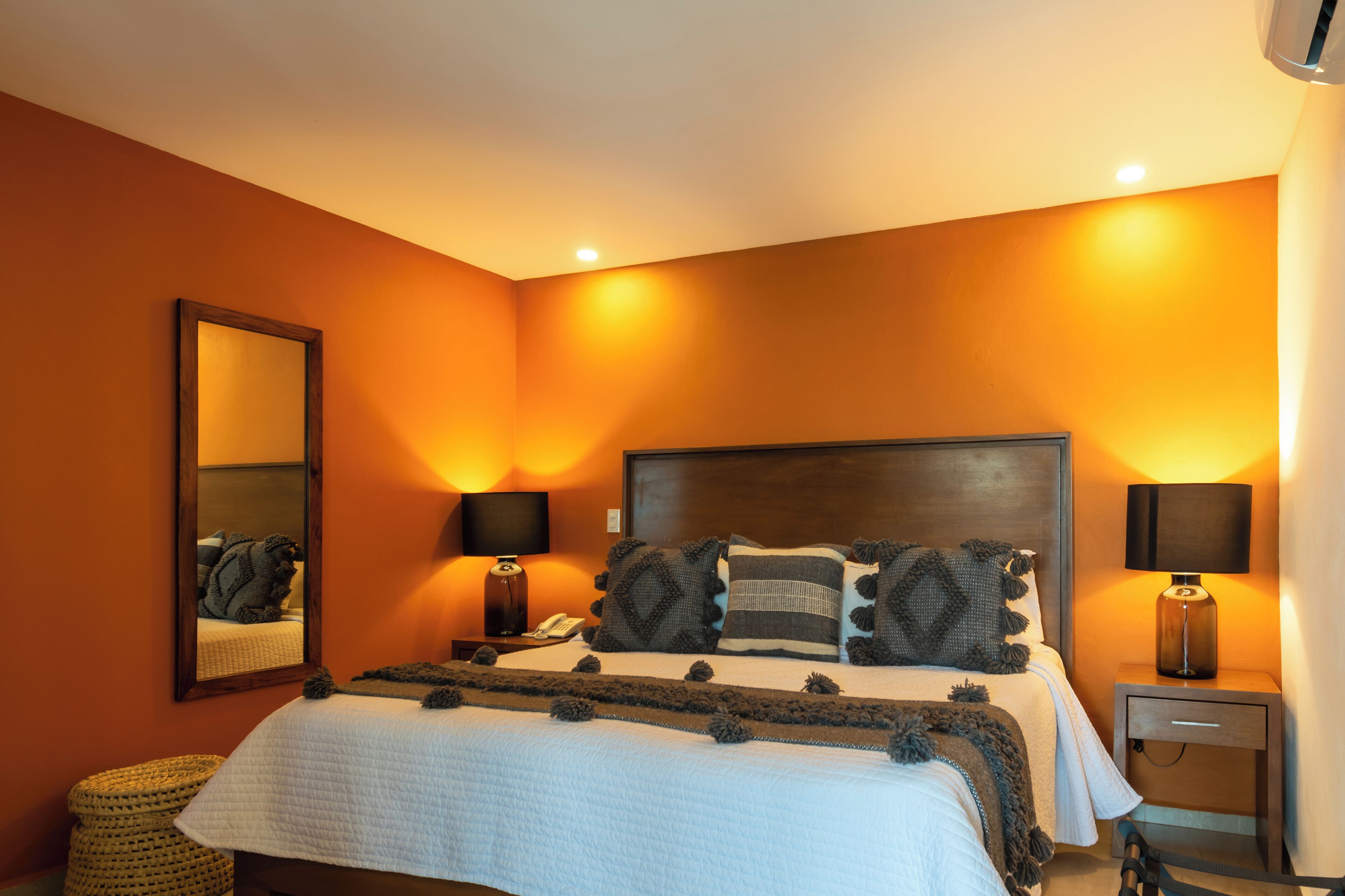 Quarto com uma cama grande, duas mesas de cabeceira com luminárias, um espelho na parede e uma cesta de palha no chão.