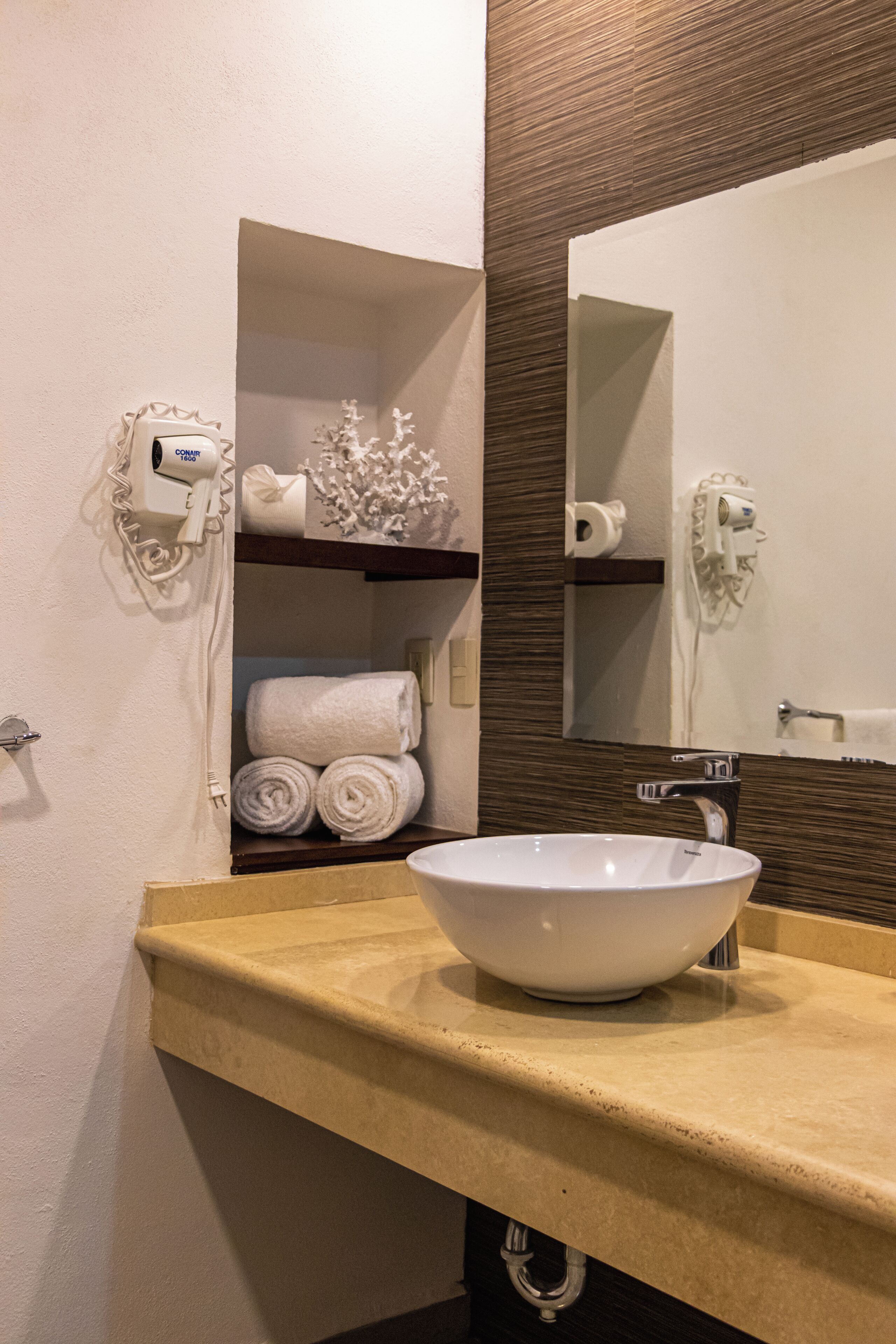 Un baño con encimera color beige, un lavabo blanco y un espejo encima. Hay toallas y un secador de pelo en un estante.