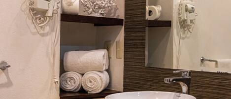 Un baño con encimera color beige, un lavabo blanco y un espejo encima. Hay toallas y un secador de pelo en un estante.