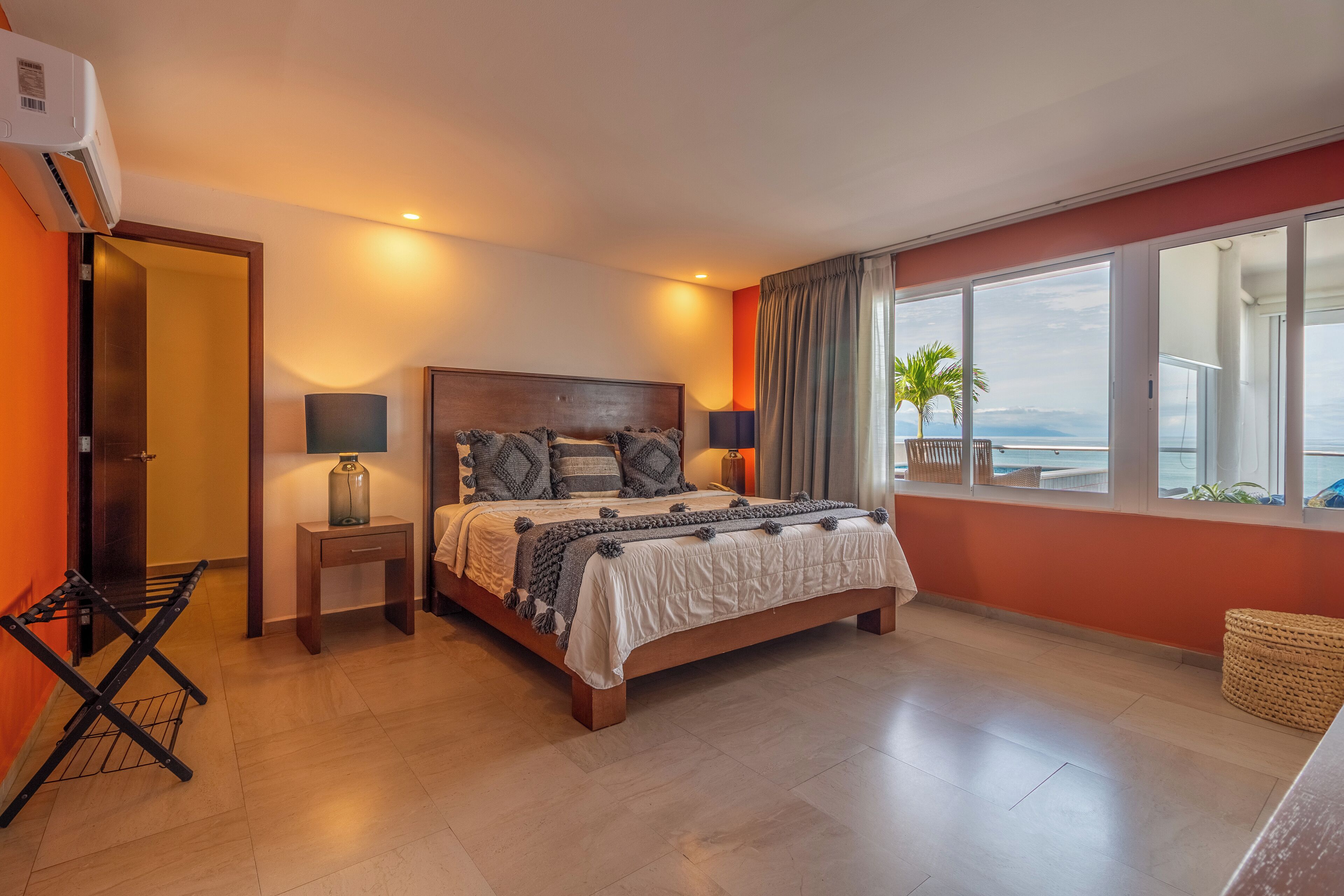 A bedroom with a bed, bedside tables, a lamp, a window with curtains, and a view of the ocean.