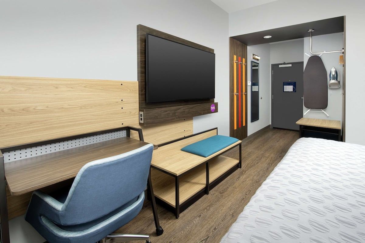 A hotel room with a wooden desk, a blue chair, a bed with a patterned duvet, a flat-screen TV mounted on the wall, and a door with a mirror and a towel rack.