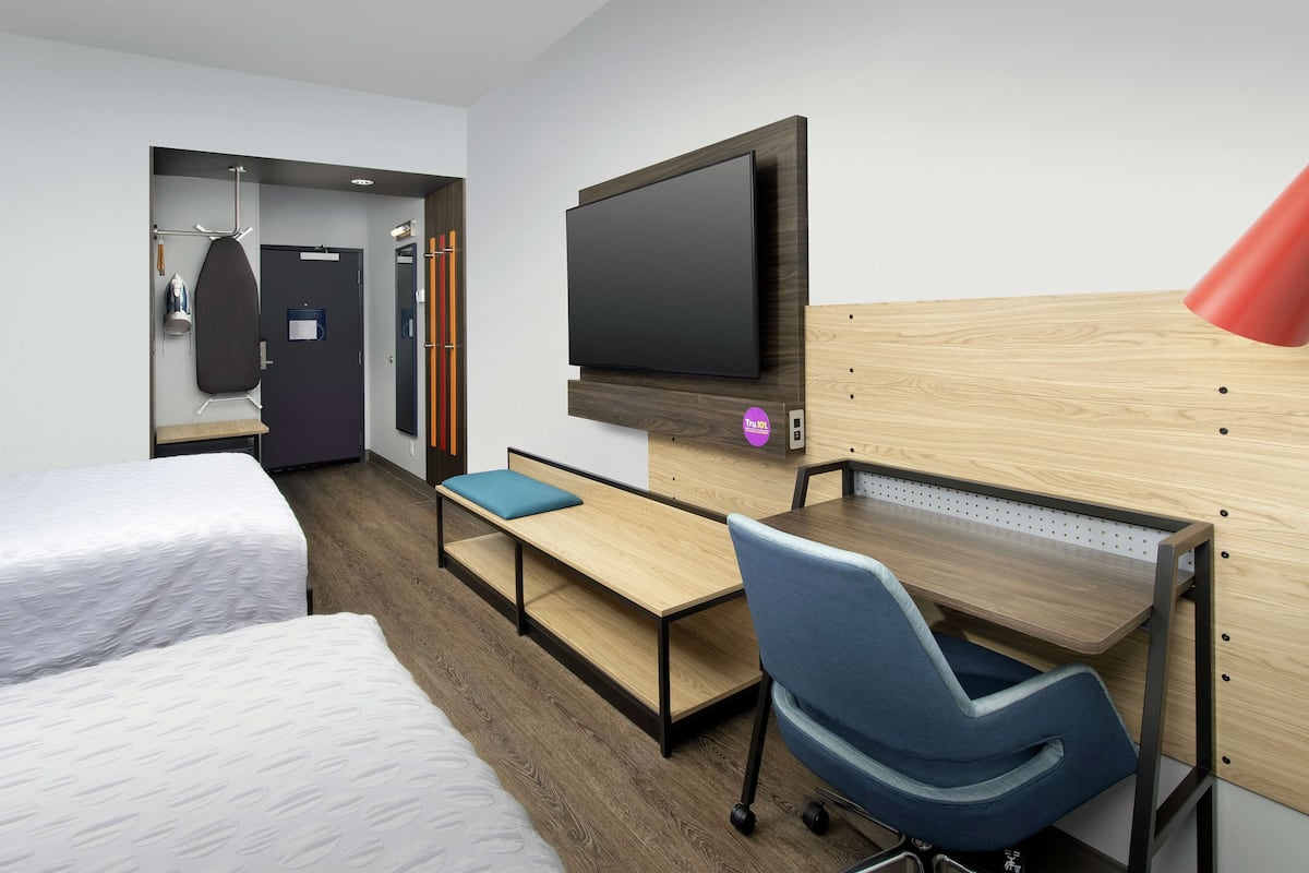 A hotel room with a bed, a desk with a chair, a TV mounted on the wall, and a red lamp.
