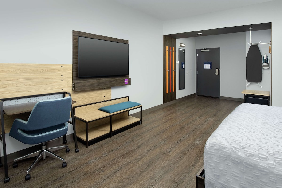A modern hotel room with a wooden desk, a blue office chair, a bed with a white mattress, a flat-screen TV mounted on the wall, and a door with a sign.