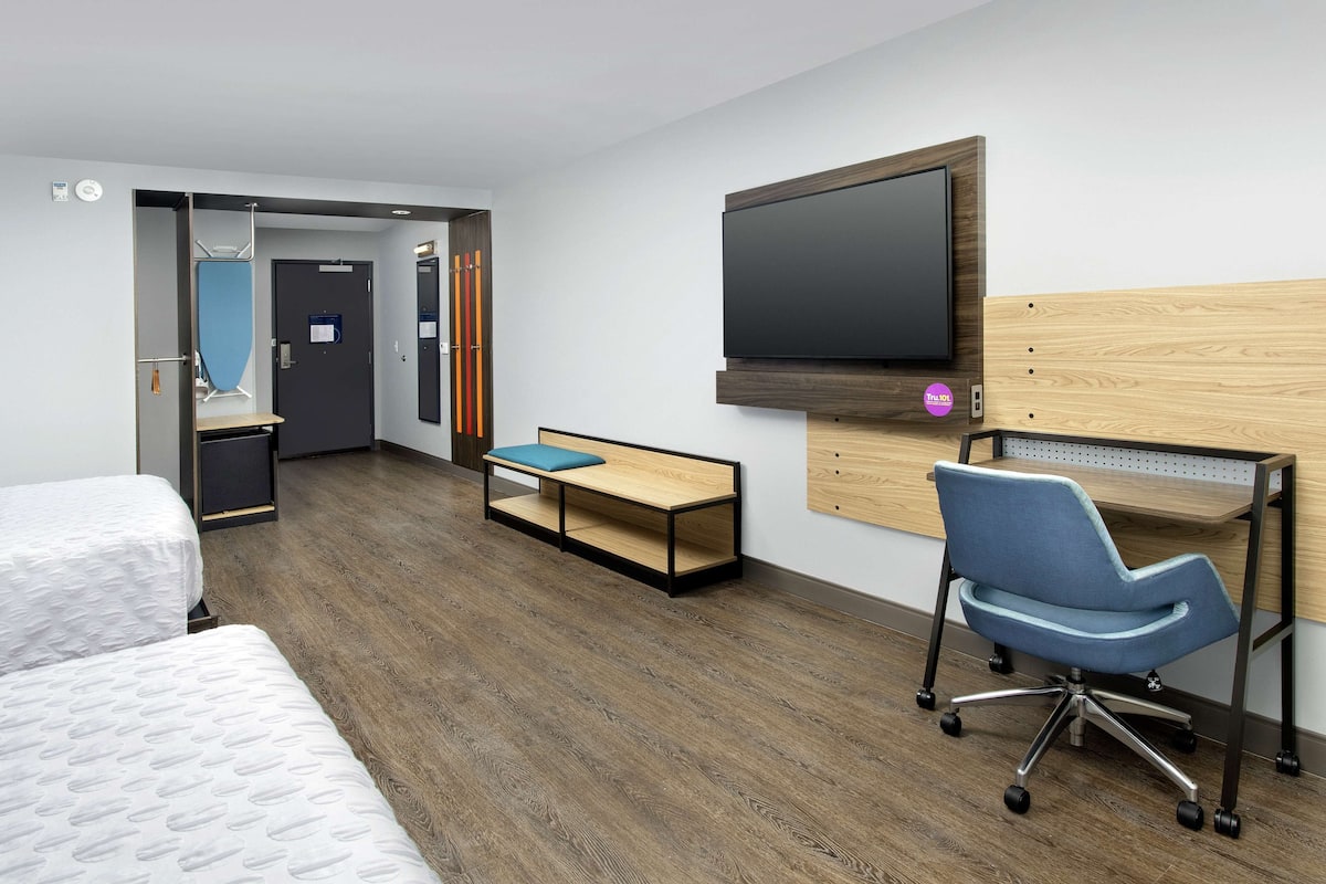 A hotel room with two beds, a desk with a chair, a TV mounted on the wall, and a small seating area.
