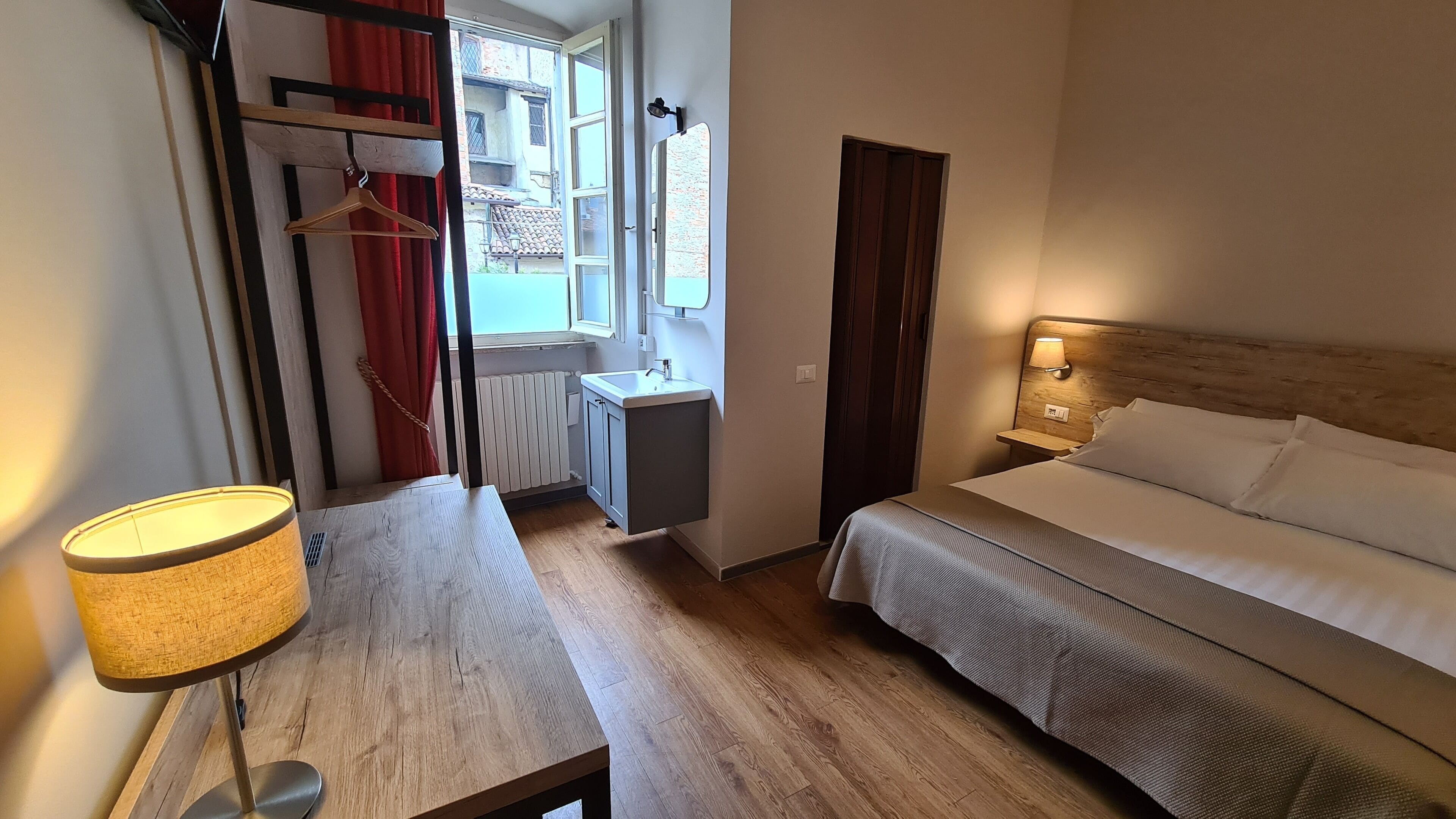 A bedroom with a bed, bedside table, lamp, and a window with a view of buildings.