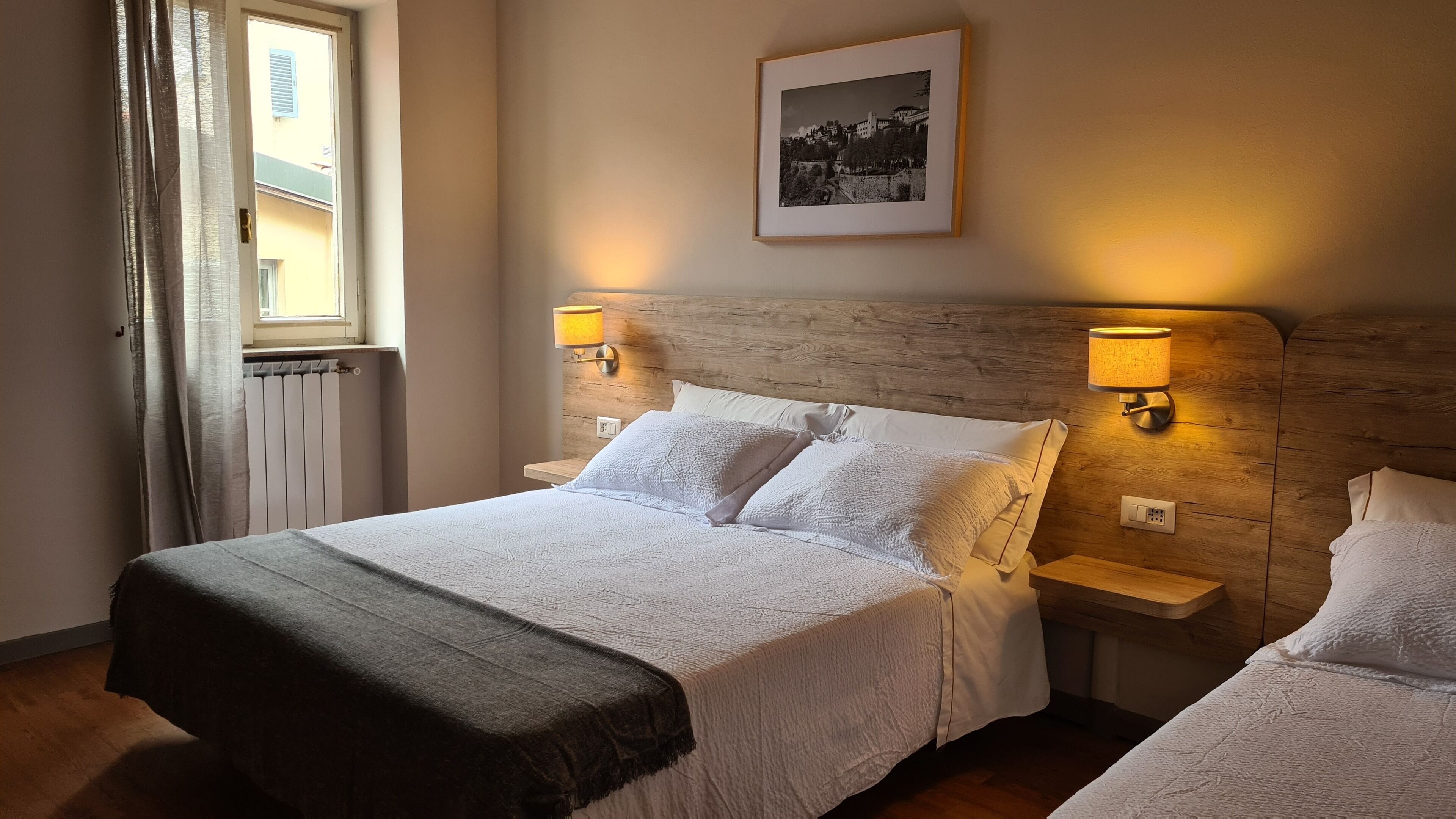 Una cama doble con cabecera de madera, dos mesitas de noche con lámparas, una ventana con cortinas y un cuadro enmarcado en la pared.