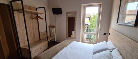A bedroom with a bed, wooden wardrobe, a mirror, a TV, and a window with a view of greenery.