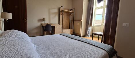 A bedroom with a large bed, a desk, a chair, a wardrobe, and a window with curtains.