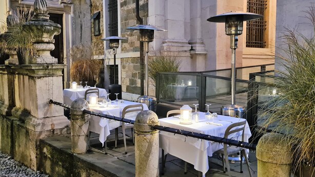 An outdoor dining area with tables set for two, each with a white tablecloth, candles, and glassware. There are three solar-powered lamps providing light. The setting is in front of a historic building with stone architecture and a wrought iron fence. - Il Sole Bergamo (Bergamo)