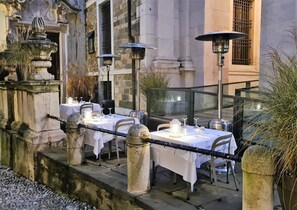An outdoor dining area with tables set for two, each with a white tablecloth, candles, and glassware. There are three solar-powered lamps providing light. The setting is in front of a historic building with stone architecture and a wrought iron fence. - Il Sole Bergamo (Bergamo)
