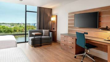 A modern hotel room with a large window, a flat-screen TV, a desk with a chair, a sofa, and a small table.