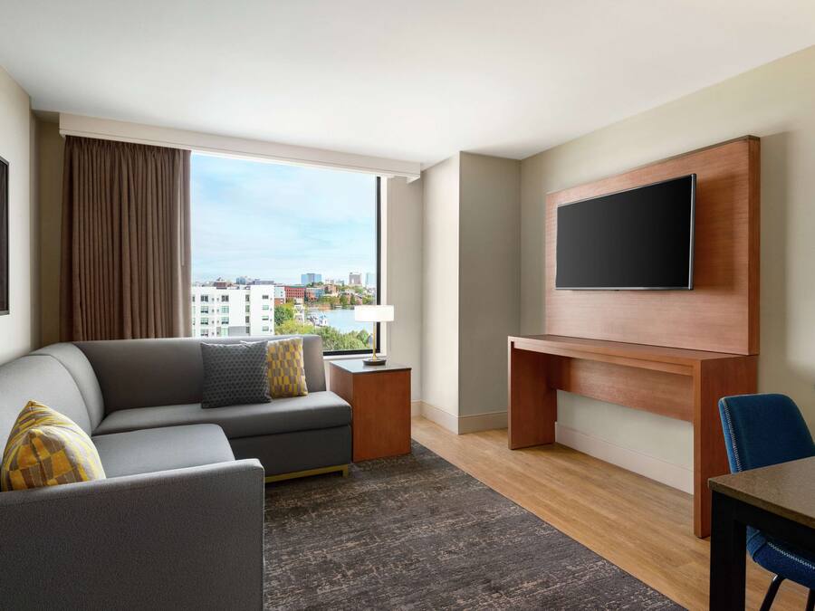 A modern hotel room with a grey sofa, a flat-screen TV mounted on a wooden wall, a dining table with blue chairs, and a large window offering a city view.