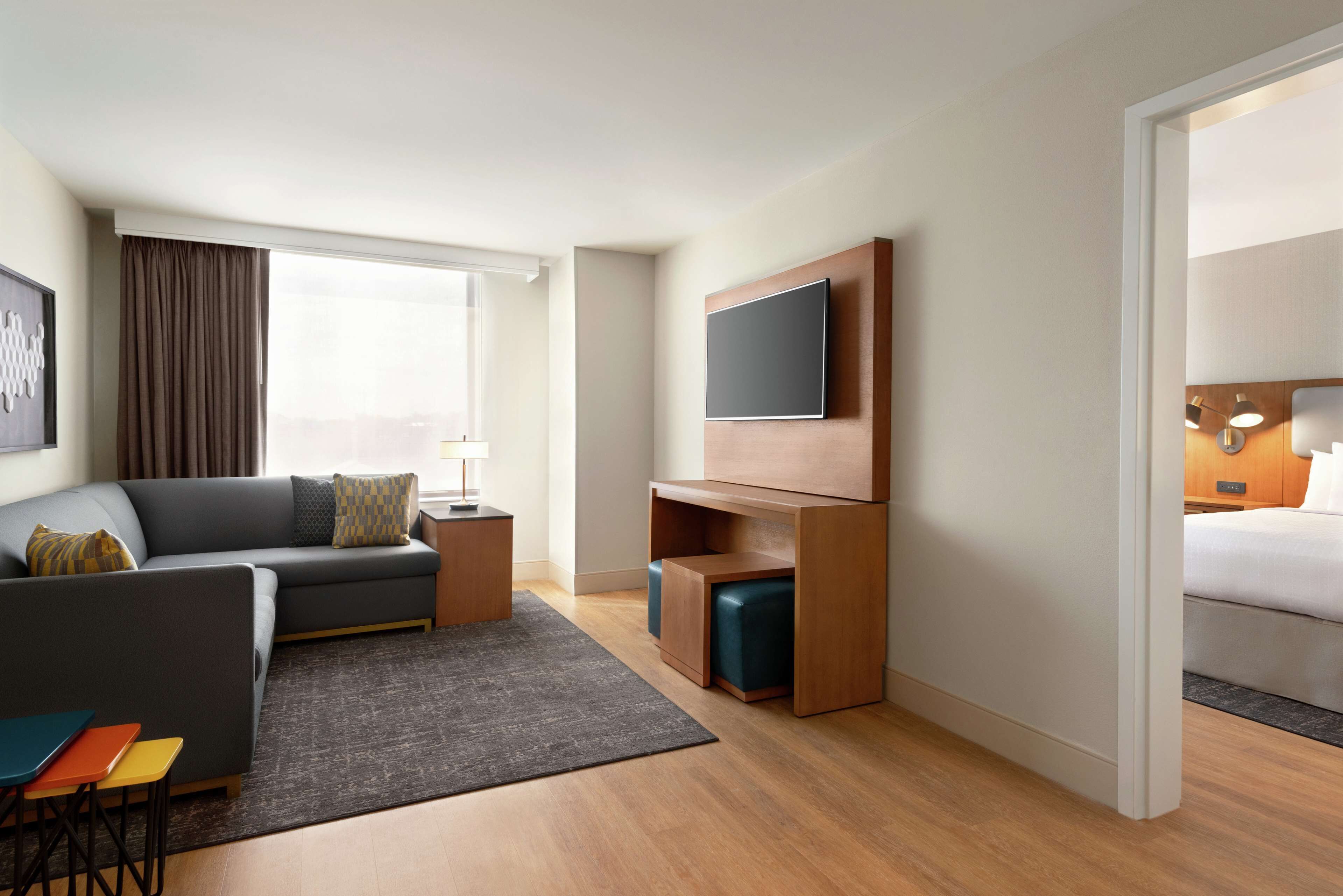 A modern hotel room with a grey sofa, a flat-screen TV mounted on a wooden panel, a small wooden side table, a grey rug, and a wooden floor.