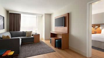 A modern hotel room with a grey sofa, a flat-screen TV mounted on a wooden panel, a small wooden side table, a grey rug, and a wooden floor.