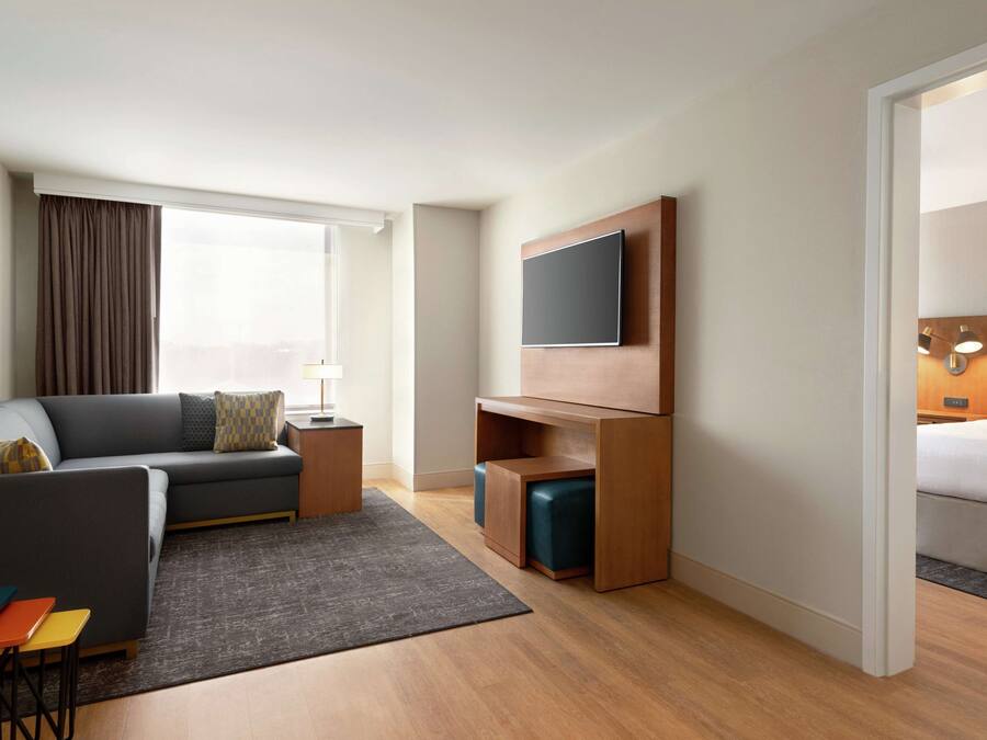 A modern hotel room with a grey sofa, a flat-screen TV mounted on a wooden panel, a small wooden side table, a grey rug, and a wooden floor.