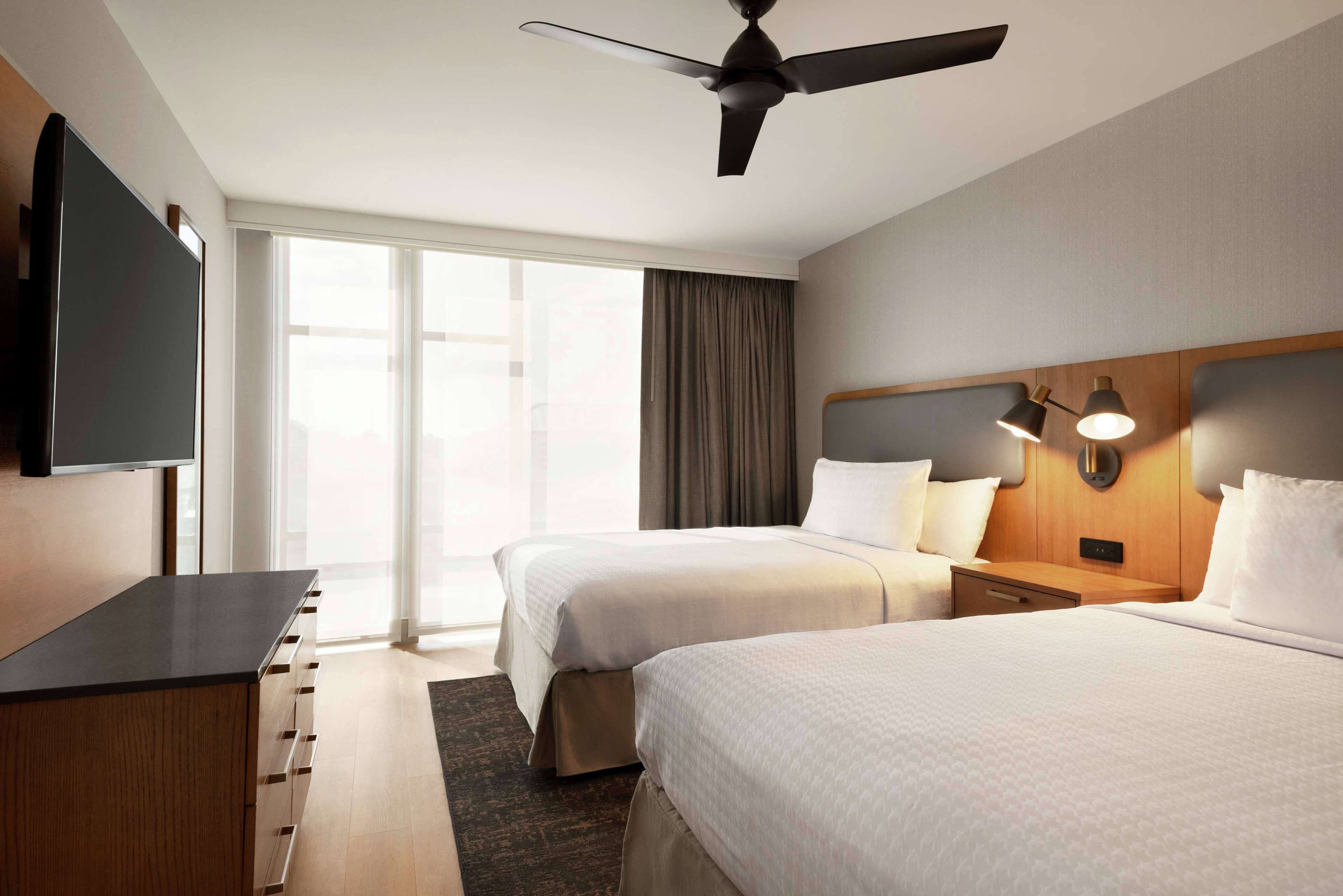 A hotel room with two beds, a flat-screen TV, a ceiling fan, and a large window with curtains.