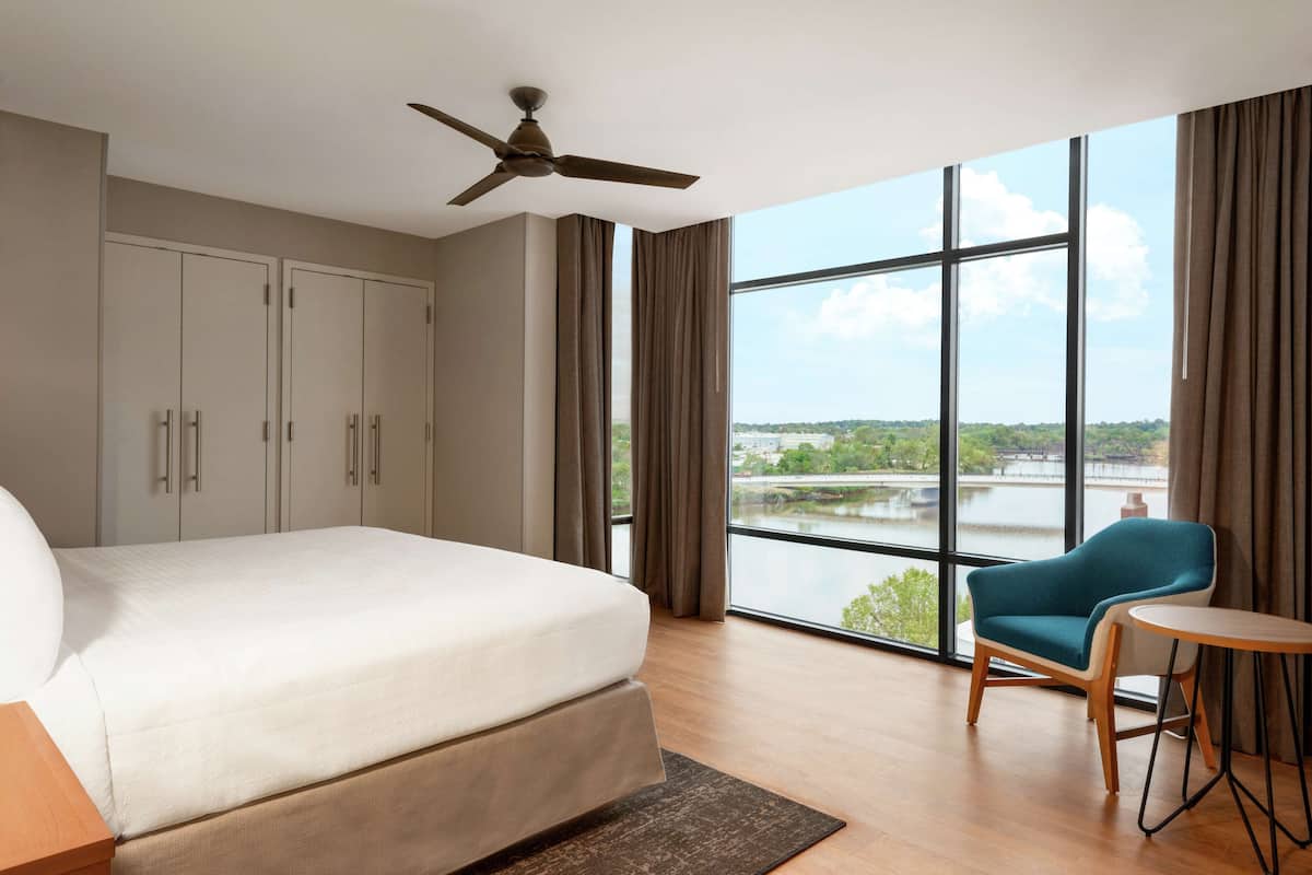 A modern hotel room with a large bed, a ceiling fan, a view of a river, and a seating area with a chair and table.
