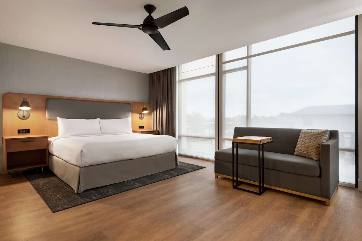 A modern hotel room with a large bed, a sofa, a ceiling fan, and a view of the city through large windows.