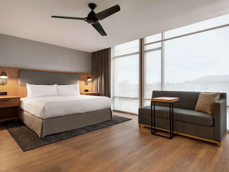 Habitación de hotel moderna con una cama grande, un sofá, un ventilador de techo y vistas de la ciudad a través de amplios ventanales.