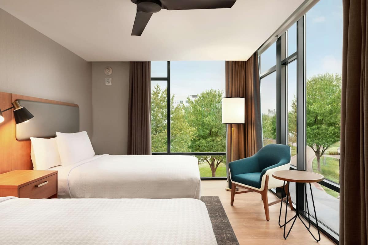 A hotel room with two beds, a chair, a small table, and large windows offering a view of trees and buildings.