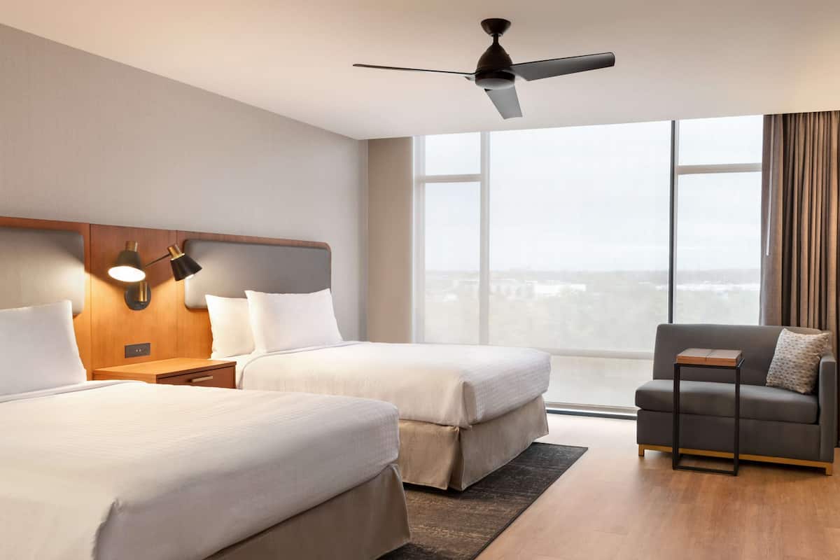 A hotel room with two beds, a ceiling fan, a nightstand with a lamp, a sofa, and a large window with a view.