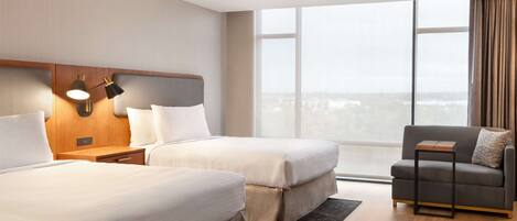 A hotel room with two beds, a ceiling fan, a nightstand with a lamp, a sofa, and a large window with a view.