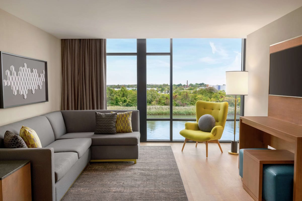A modern living room with a grey sectional sofa, a yellow armchair, and a view of greenery and buildings through large windows.