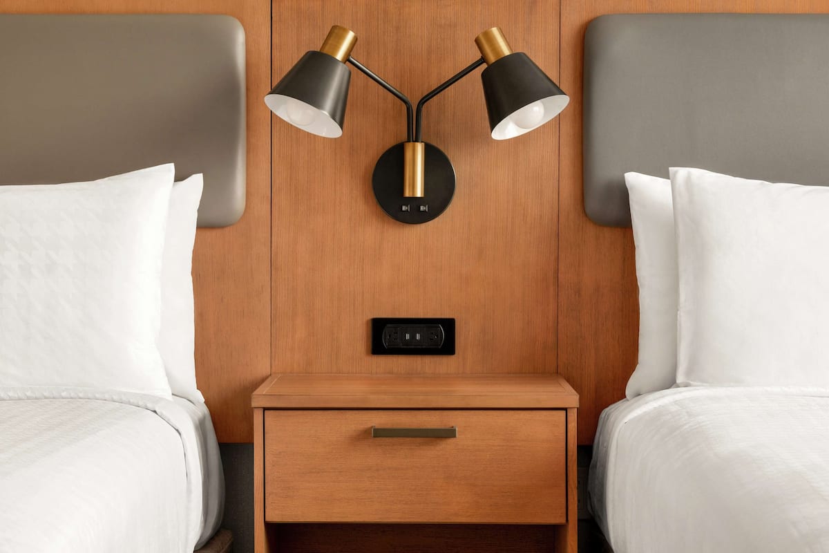 A hotel room with two beds, a wooden nightstand with a drawer, and a wall-mounted lamp.