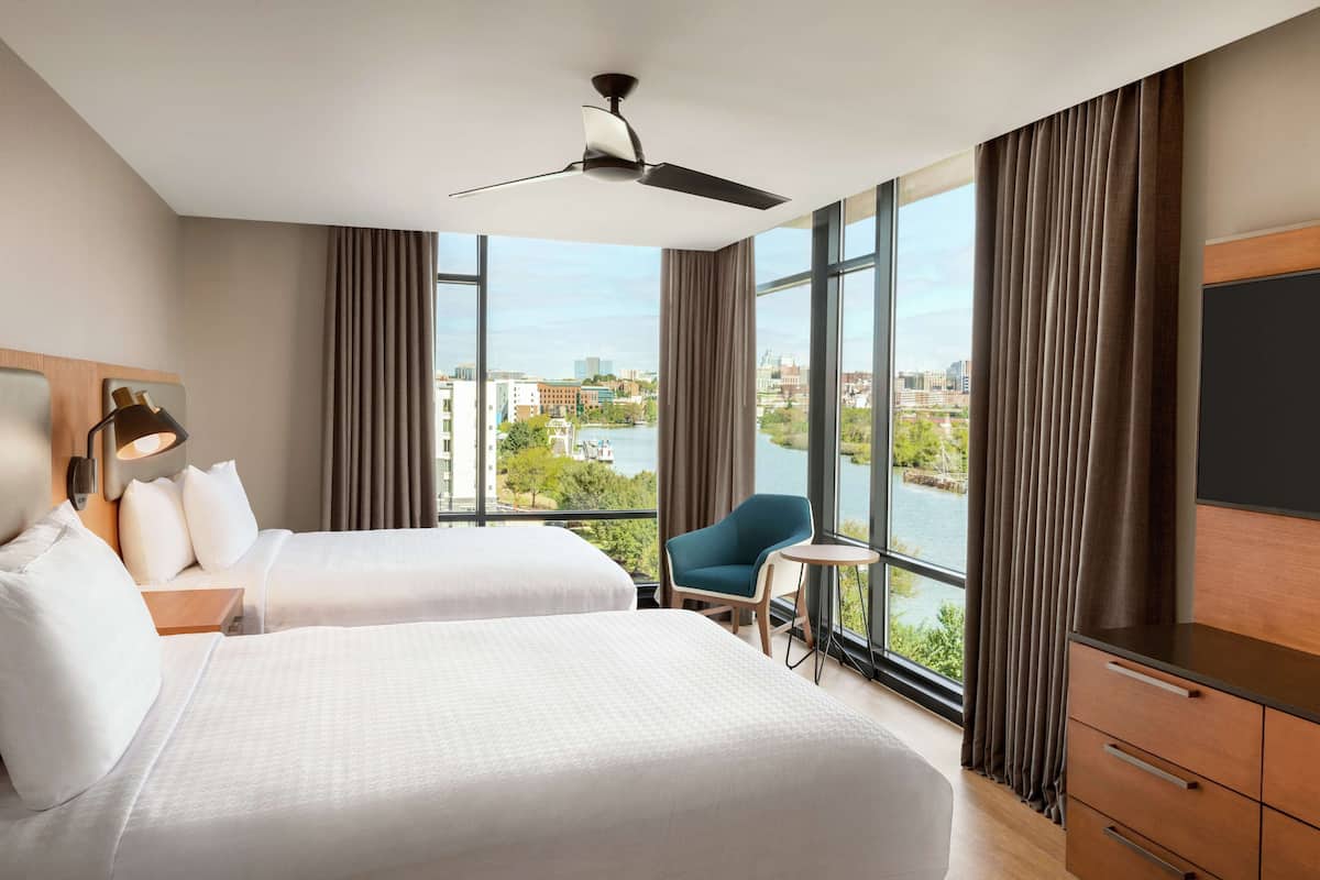 A hotel room with two beds, a desk, a chair, a TV, and a view of a city and river.