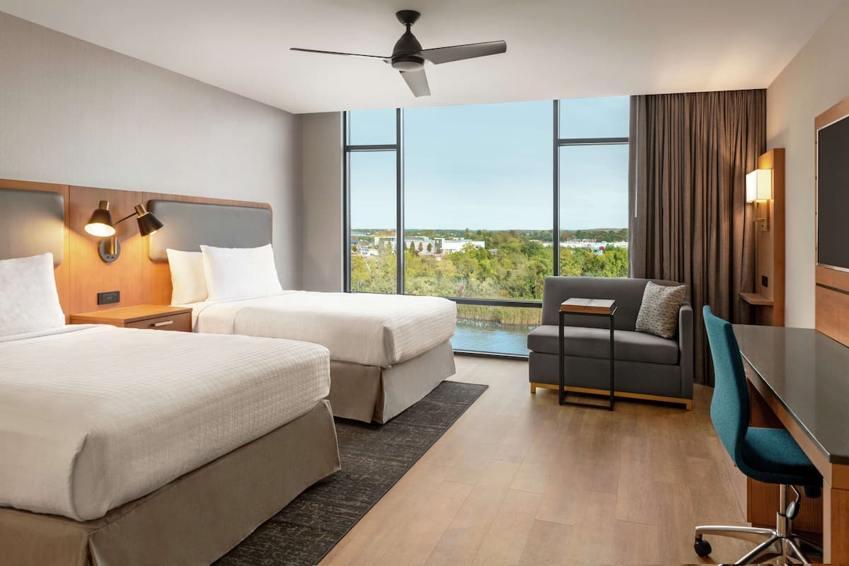 A hotel room with two beds, a desk, a chair, a ceiling fan, and large windows offering a city view.