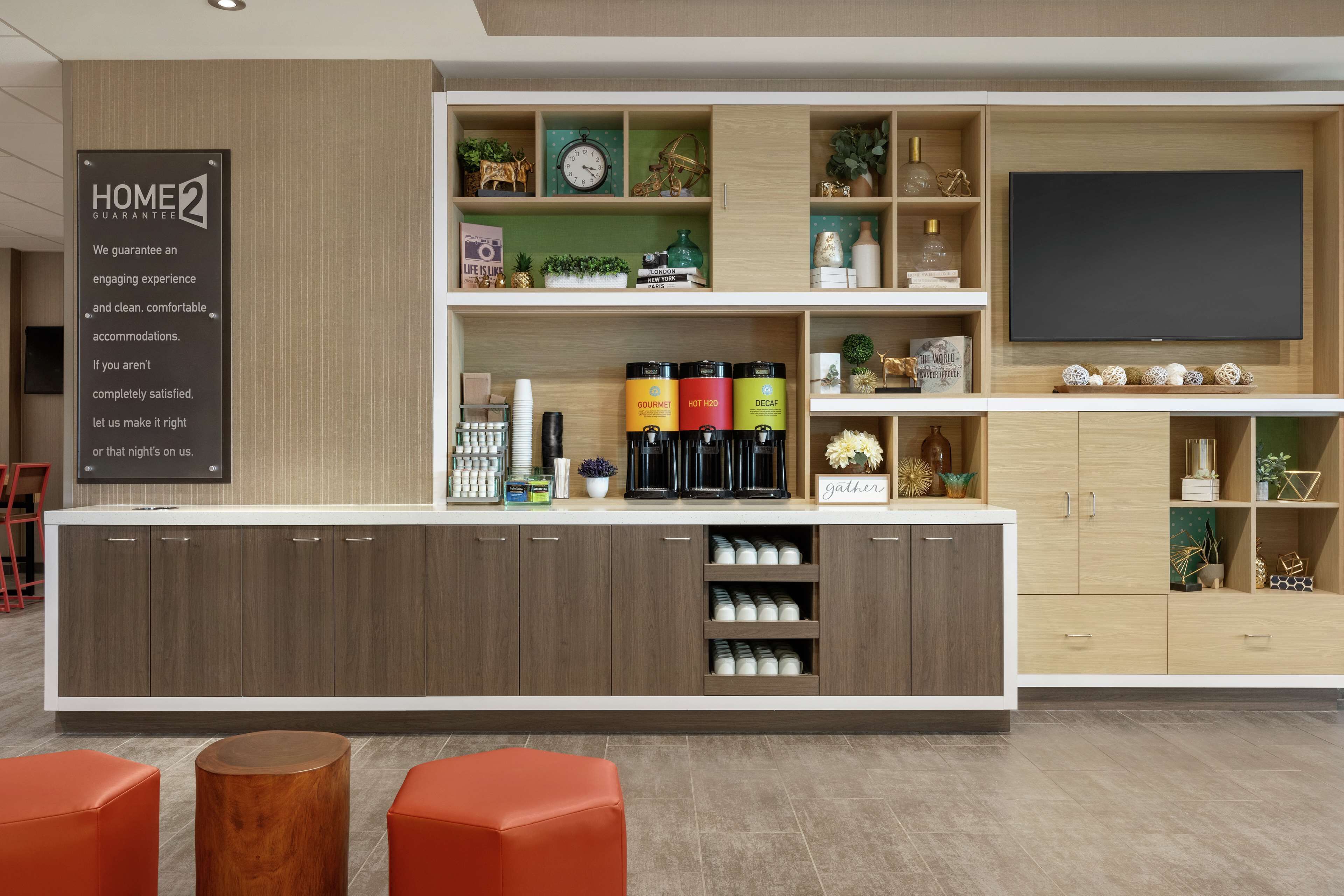 A modern hotel reception area with a wooden counter, a large flat-screen TV, and a sign stating "HOME2 GUARANTEE".