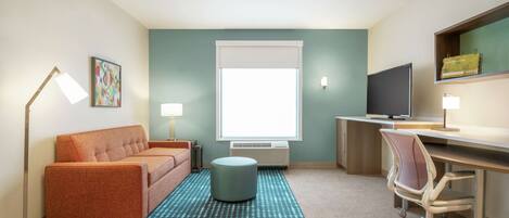 A compact hotel room with a sofa, a desk, a computer, a television, and a window with blinds.