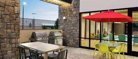 Un patio al aire libre con un muro de piedra, una parrilla, una mesa con sillas y un paraguas rojo.