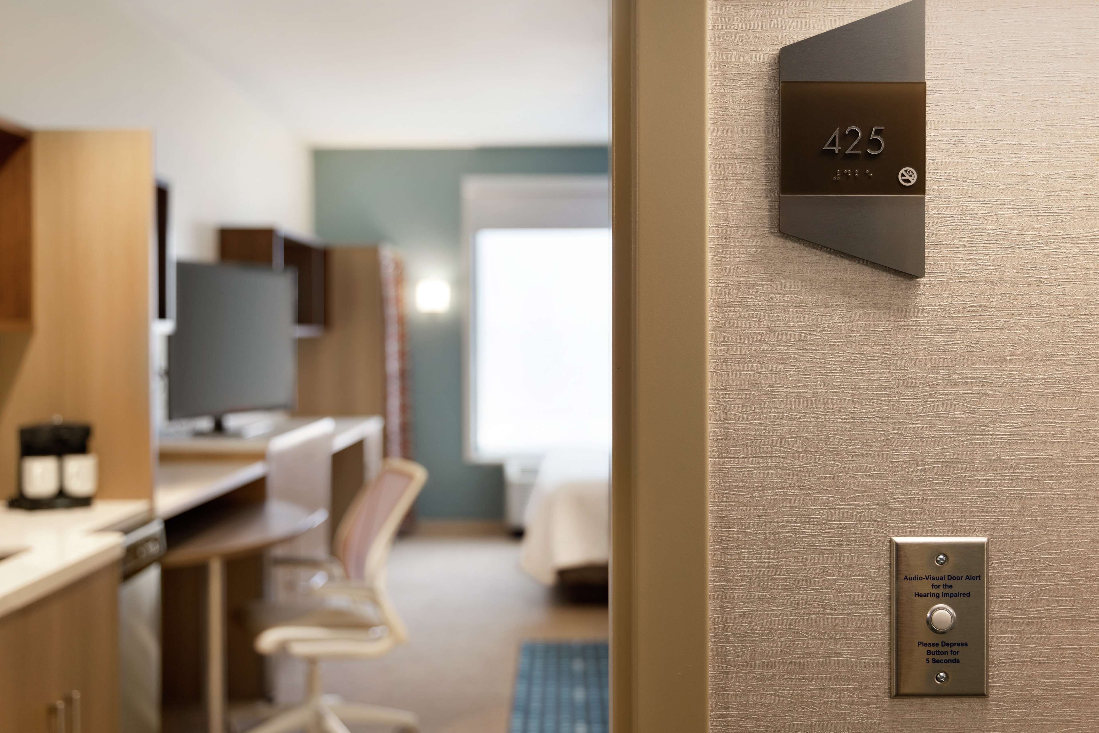 Una habitación de hotel moderna con una pantalla digital que muestra el número de habitación 425, un escritorio empotrado con una computadora, y un área de descanso con una silla.